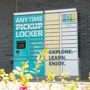 Image shows a four-column metal locker unit with the words "ANYTIME PICKUP LOCKER" and "EXPLORE. LEARN. ENJOY."